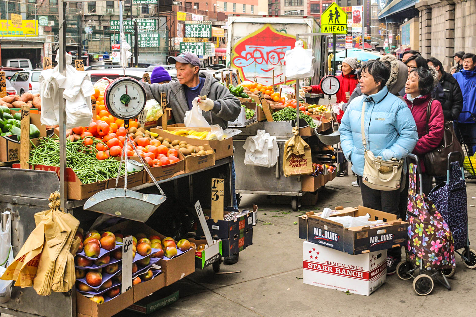 A colorful photo of a produce stand on a crowded street with a line of people waiting to shop.