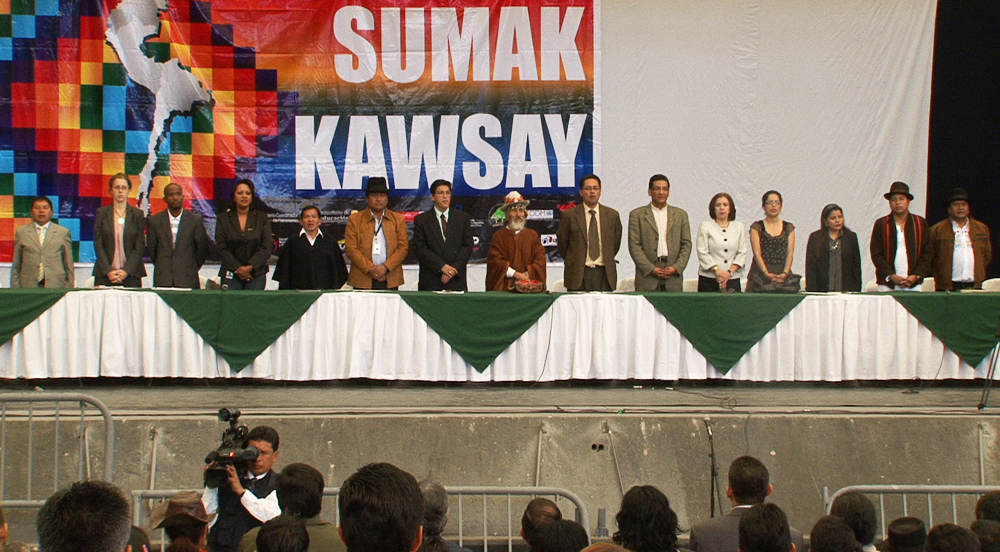 15 men and women stand on a long stage behind a conference table, with an audience in front of them. Above them is a banner with the words, :"Sumak Kawsay."