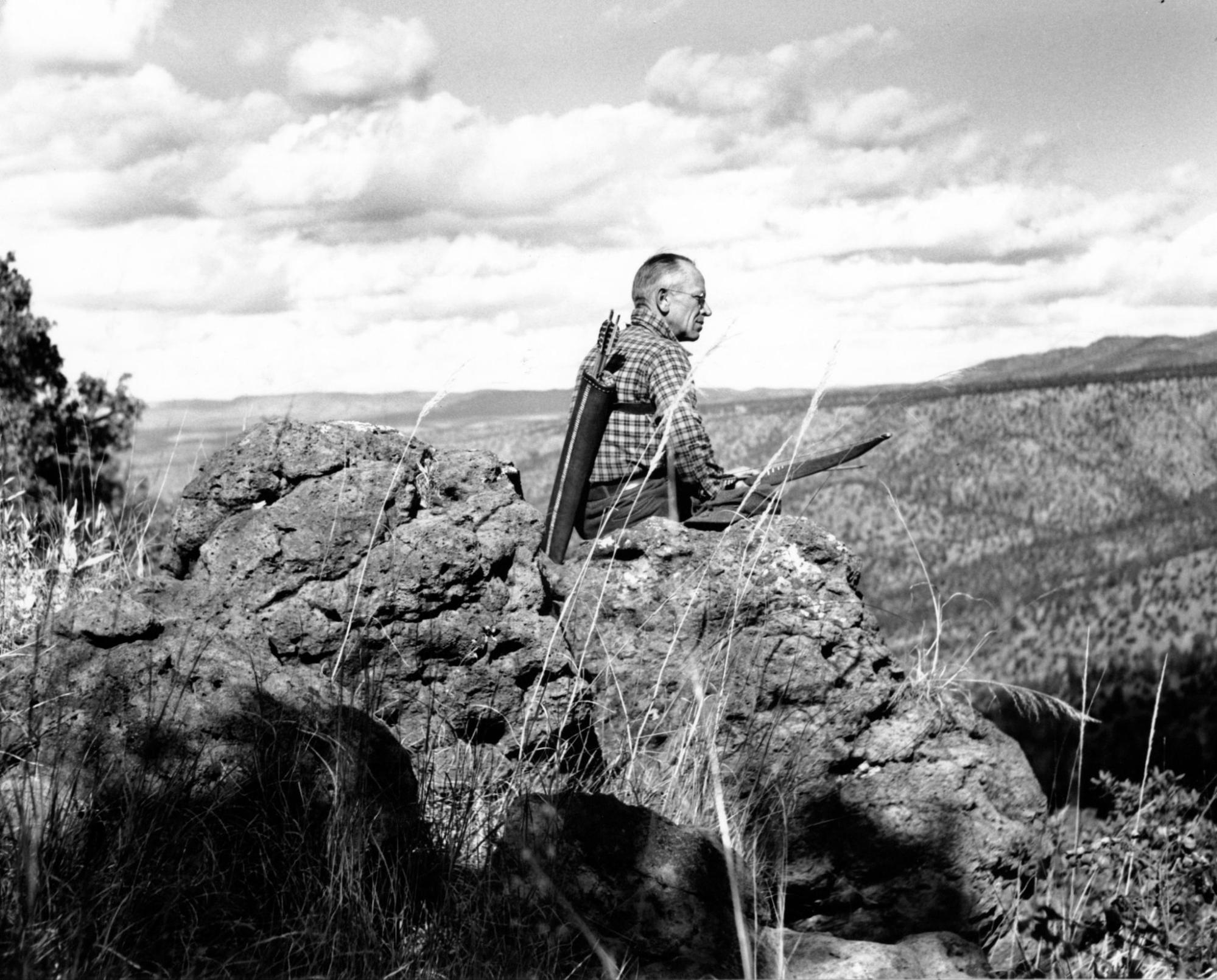 a black and white photo of man with light skin sitting at an overlook holding a quiver and bow.