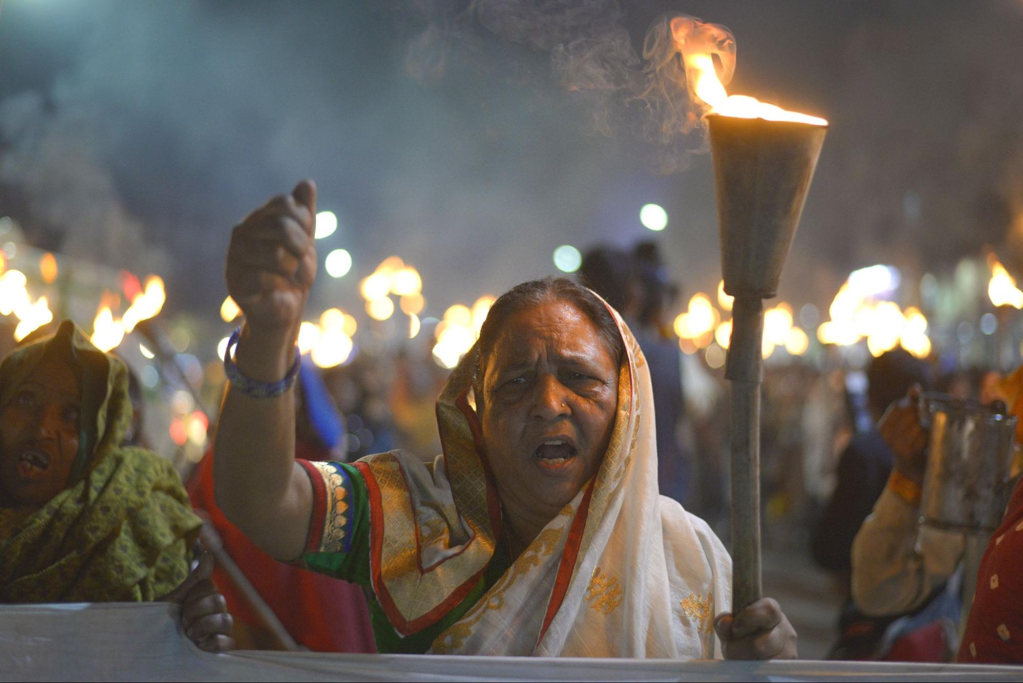 a woman wearing dress of india stands with her fist in the air holding a lit torch. Many other people carrying torches stand behind her.