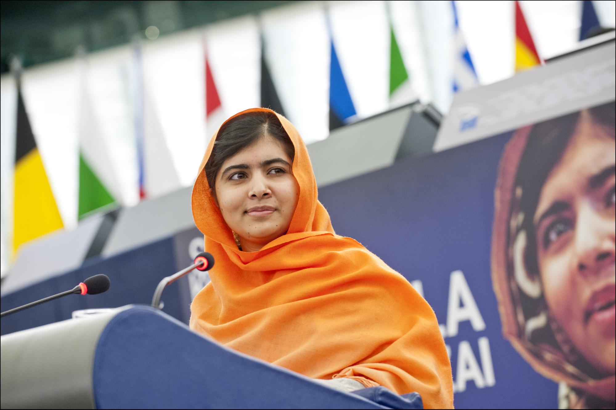 Malala Yousafzaï wearing a bright orange head scarf standing at a podium in front of her image on a billboard.