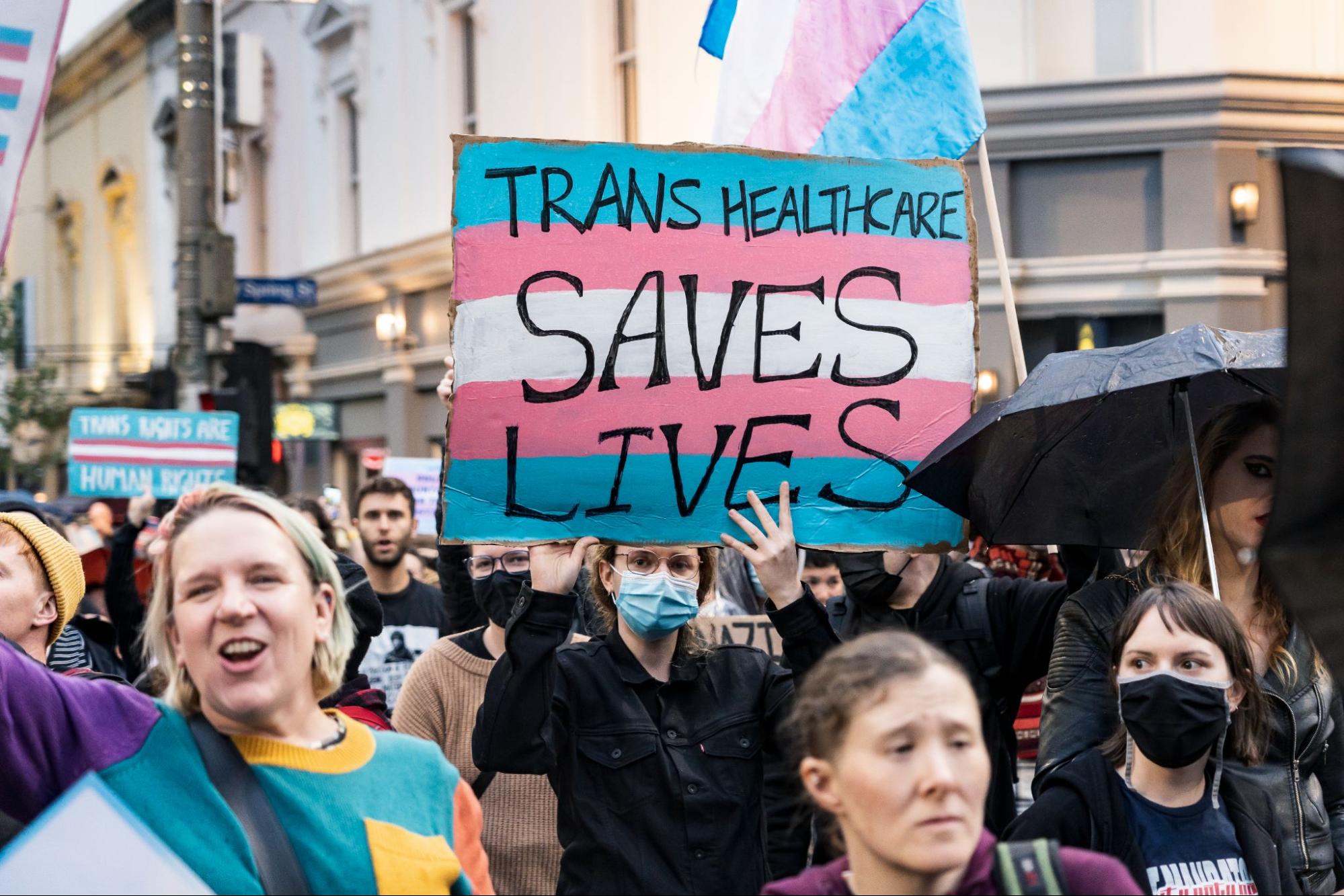 a large of people with light skin walk in the street; some with face masks. one holds a sign that reads, trans healthcare saves lives.
