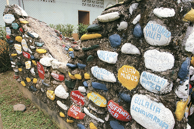 A wall is built from dirt various round stones painted red, yellow, blue, and white, with names painted on them.