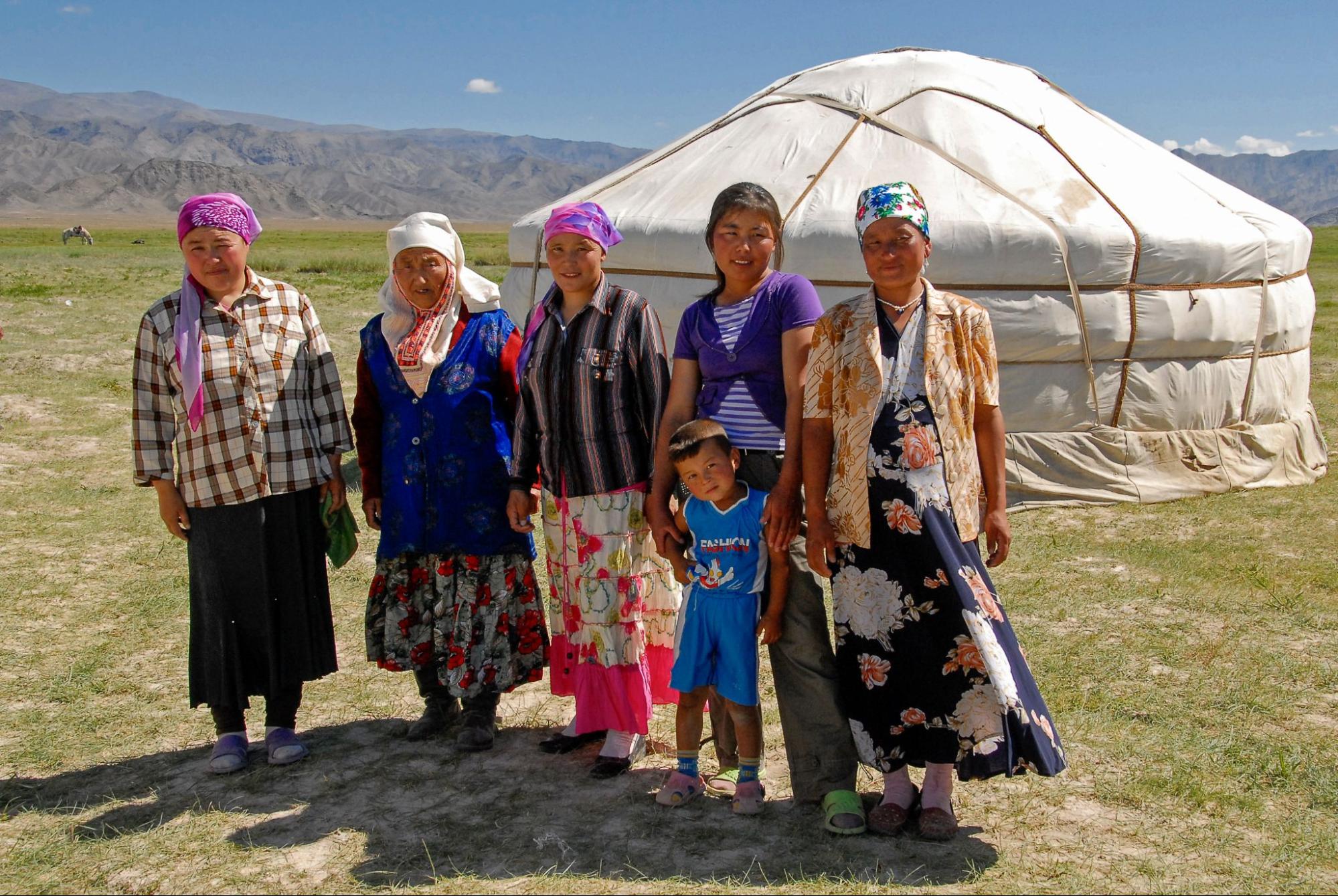 three women of different ages, and a child stand in front of a yurt with a wide landscape view behind them.