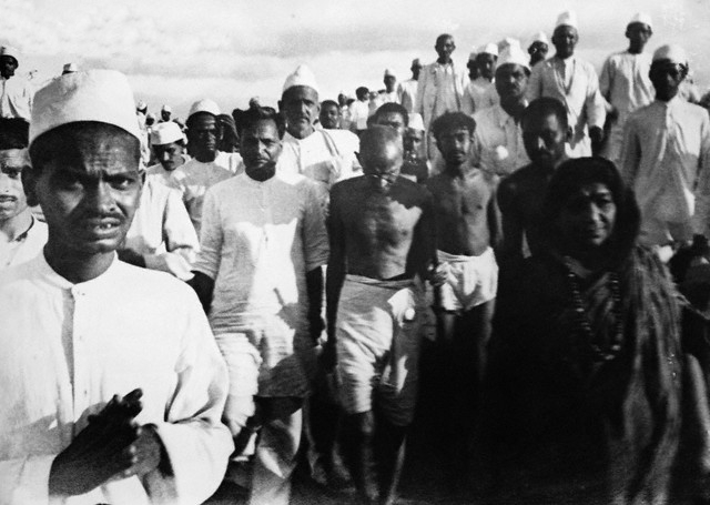 a black and white photo of Gandhi walking surrounded by many people; all are walking toward the camera.