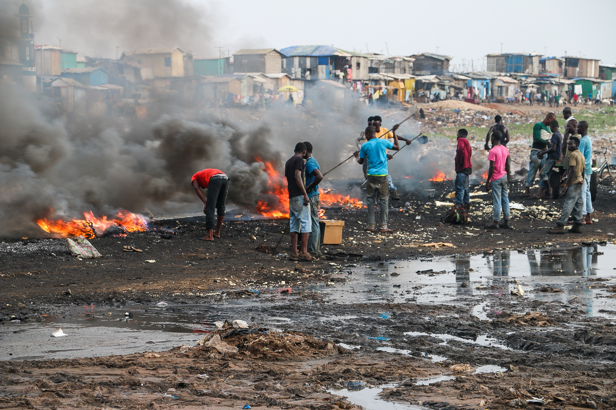 a small group of people stand in a muddy area, with fires nearby that emit very dark smoke. behind them are very low income, informal settlements