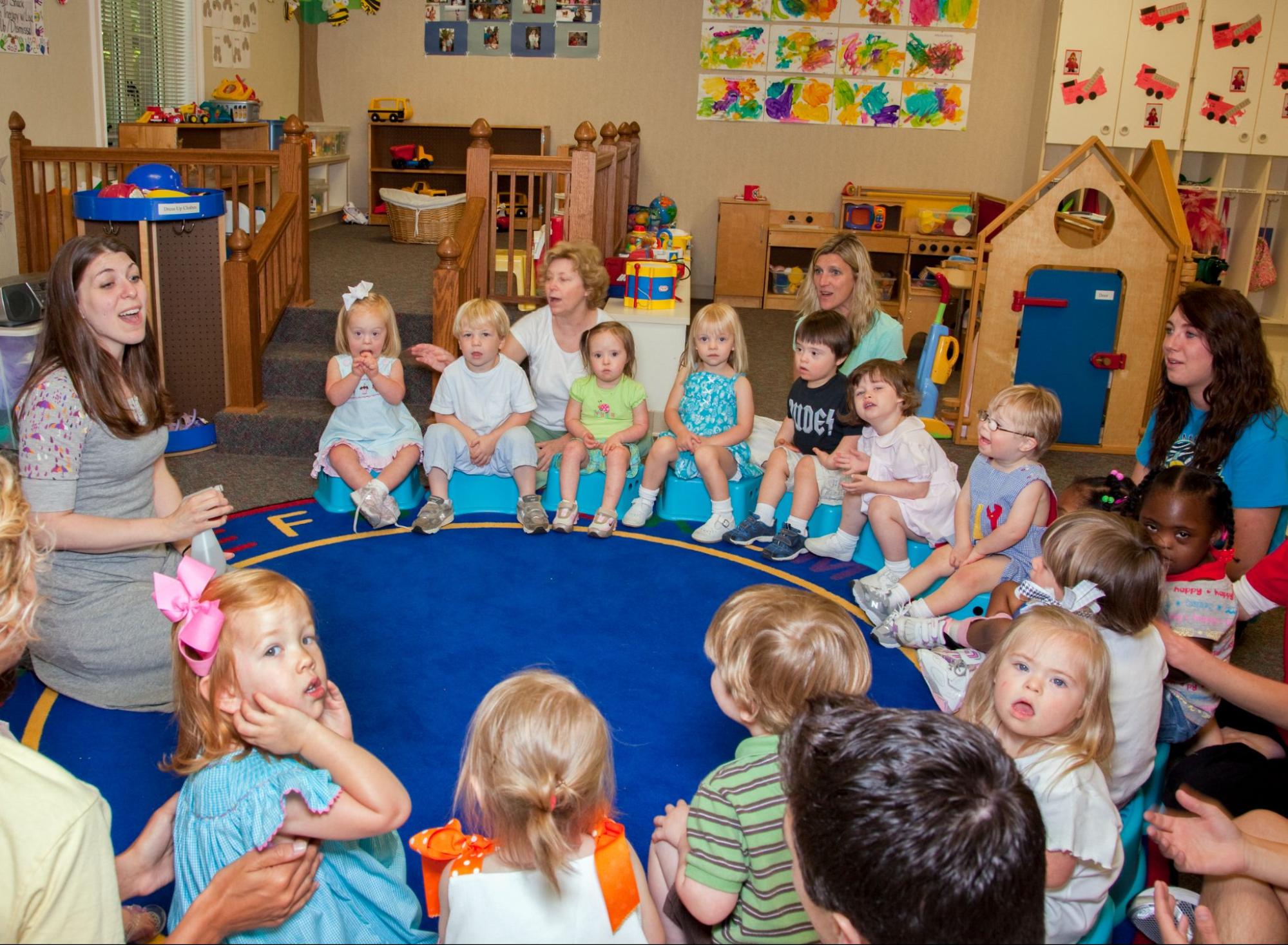 A group of young children sitting in a circle in a brightly decorated classroom with a teacher and multiple adults.