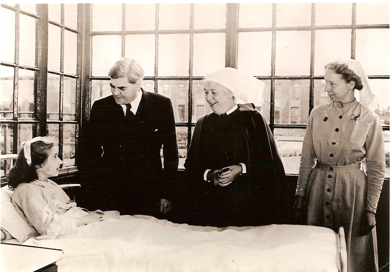 a black and white photograph of four light-skinned people. A girl in a bed, visited by a man in a suit, and woman in a nun's habit, and woman in a nurse's uniform.