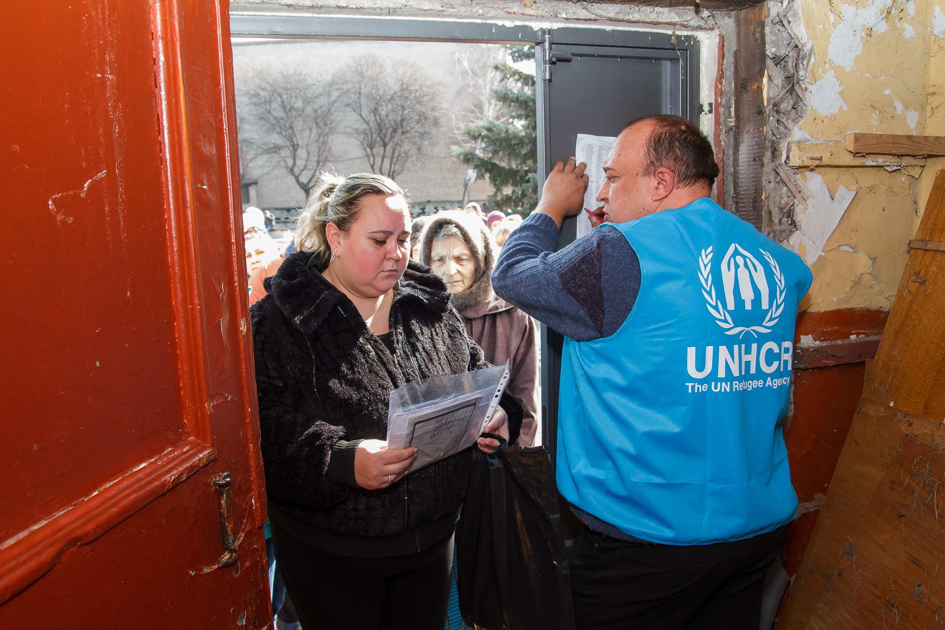 photo of a man with a United Nations Human Refugee Agency vest standing in the doorway of a building with peeling paint. At the threshold is a woman holding papers, and others behind her.