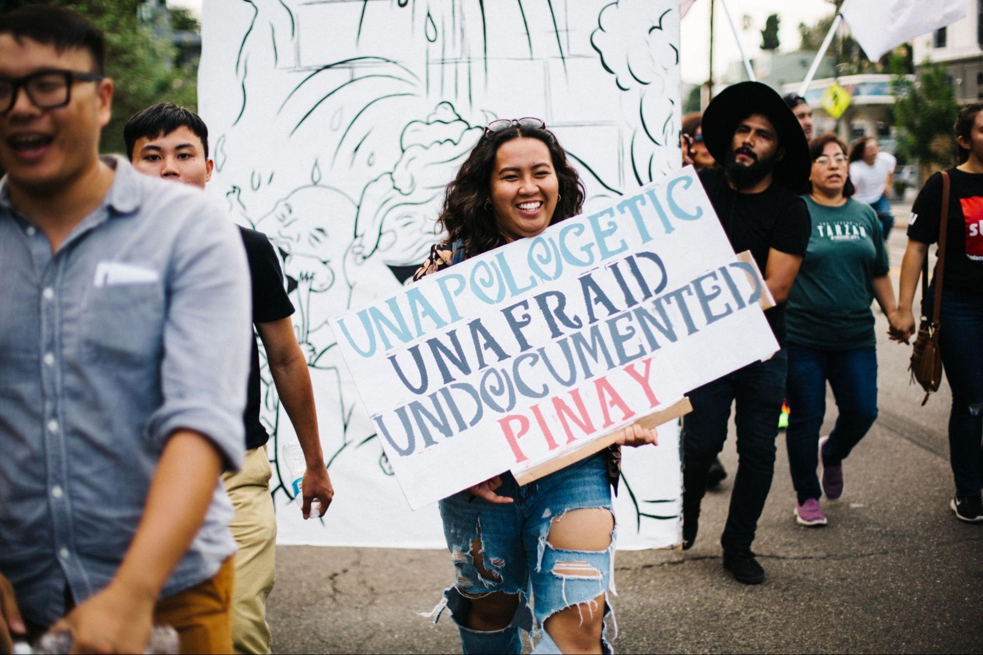 A smiling protester with curly long hair and brown skin carries a sign that reads "Unapologetic, Unafraid, Undocumented Pinay."