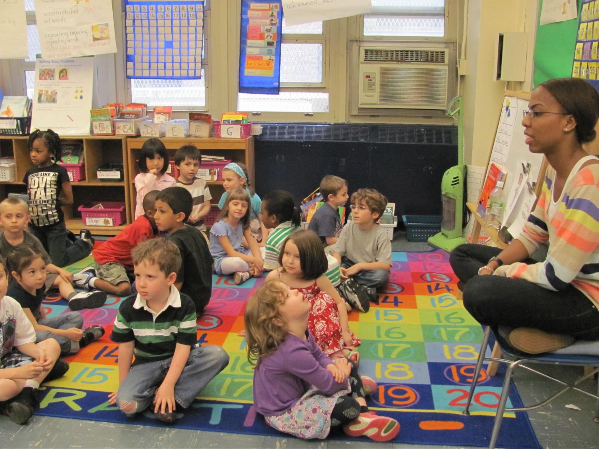 A Black female teacher sits at the front of a class with 16 young children of various ethnicities sitting on floor,