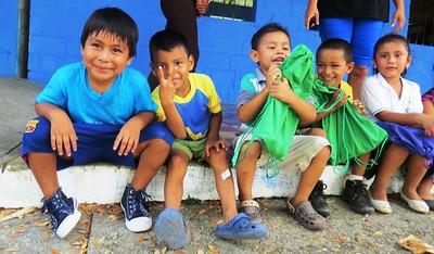 Five children of color sit next to each other on a curb, smiling