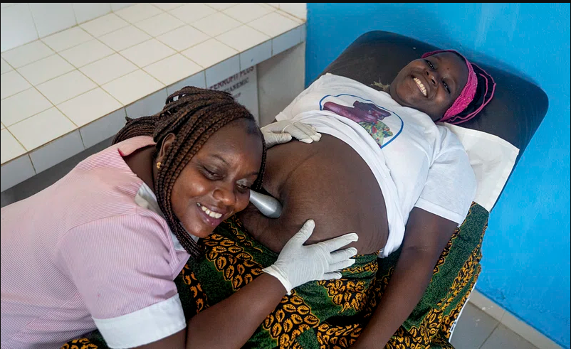 An female African healthcare provider uses a tool to listen to the pregnant African mother's belly.