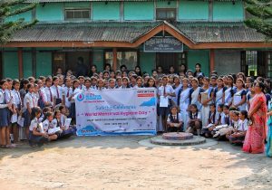 A group of people in school uniforms gather around a banner that says "Satirtha celebrates World Menstrual Hygiene Day."