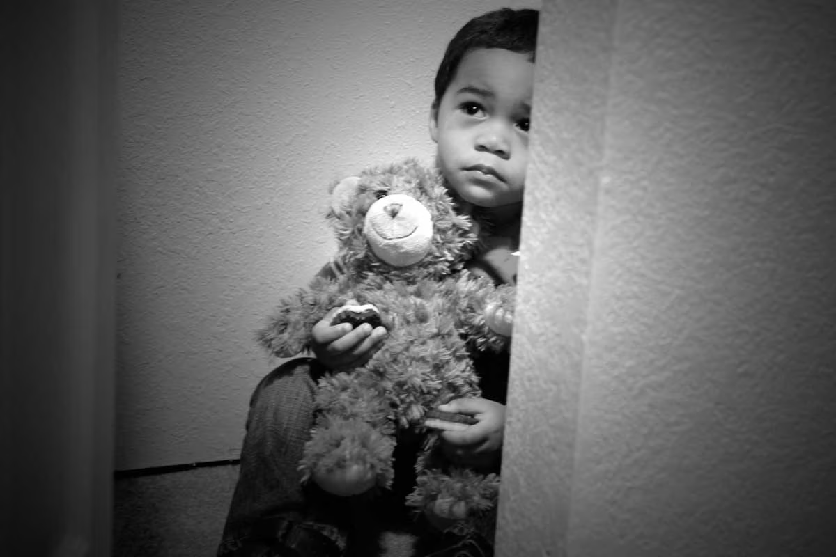 A Black make child seemingly hides behind a wall holding stuffed teddy bear