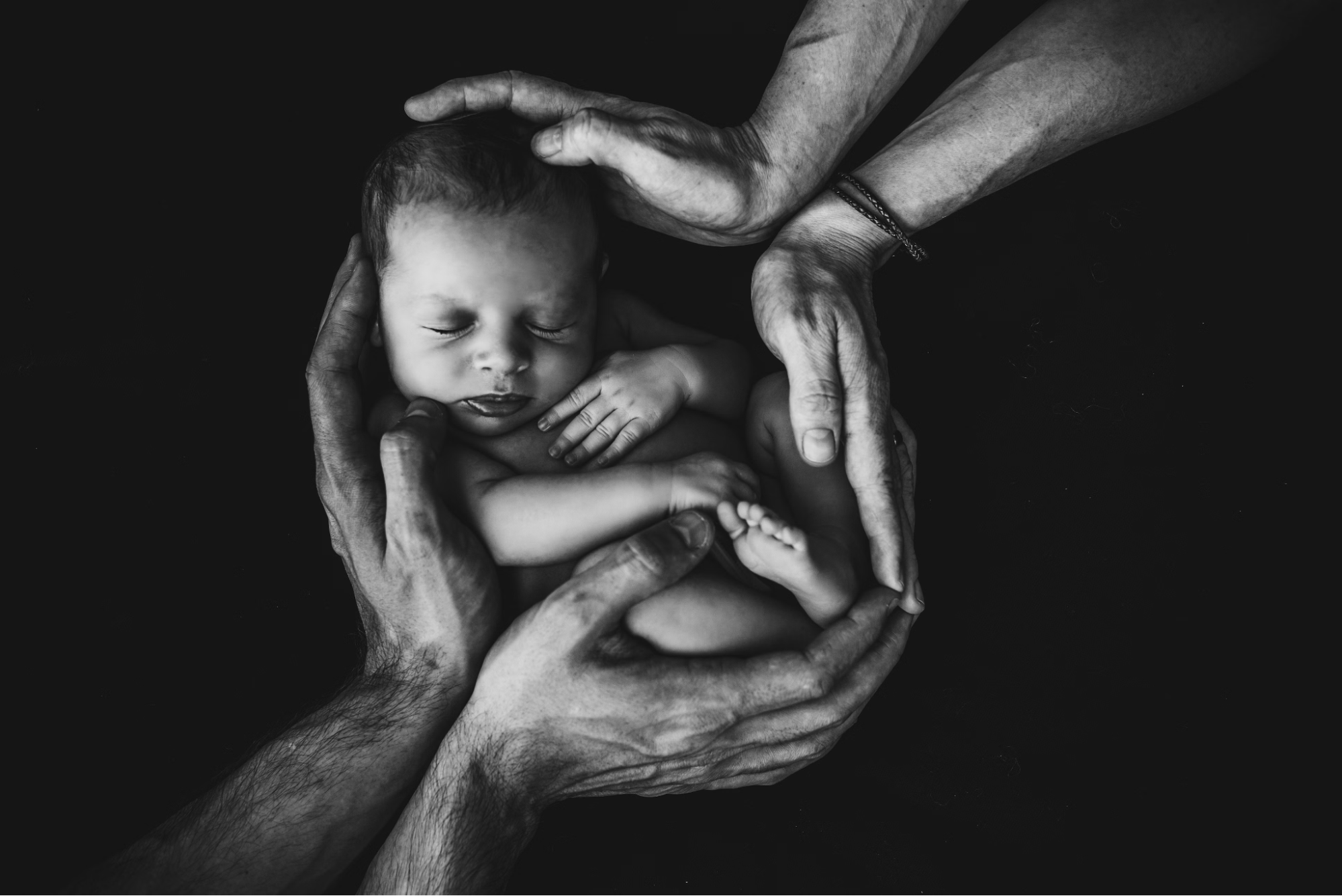 A newborn baby is held by 2 pairs of hands, possibly of different ethnicities