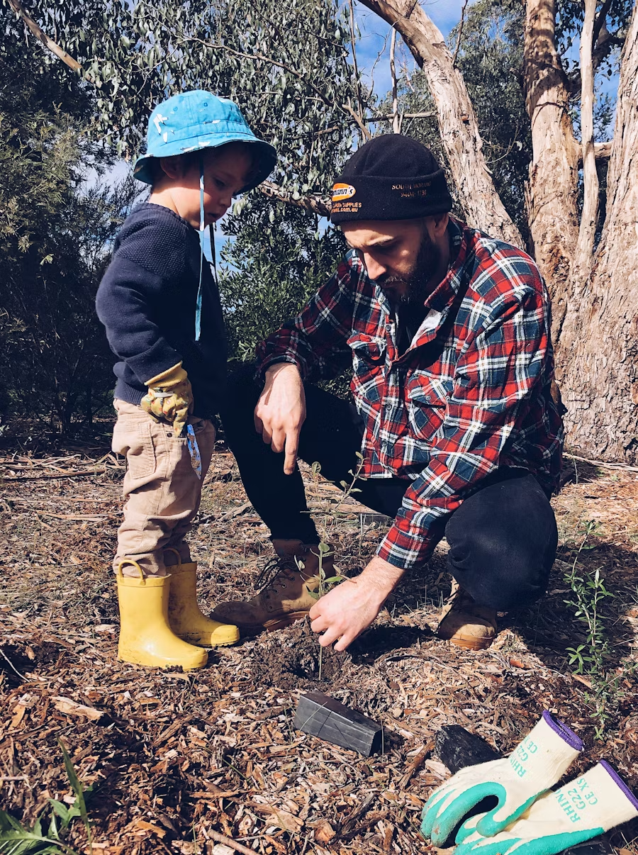 A White, male toddler stands next to a White man, demonstrating planting a small tree