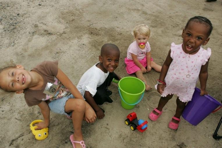 A group of Black and White children play with toys in the sand and smile up at the camera