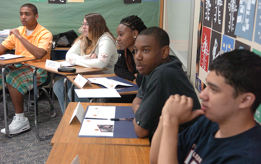 Adolescent students with light and dark skin tones sit at desks in a classroom.