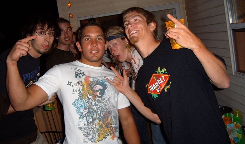 A group of teens with light and medium dark skin tones and facial hair gesture towards camera. One holds a full plastic cup.