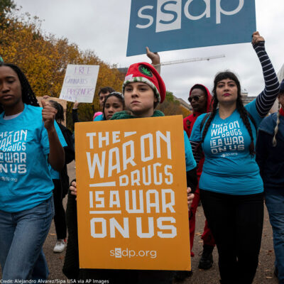 A group of individuals marching down a street protesting. In the front, a person holds a sign that says "the war on drugs is a war on us"