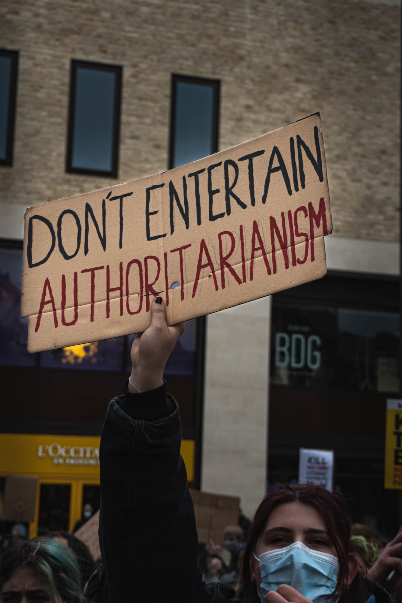 A person wearing a face mask holds a sign up in the air that says, "don't entertain authoritarianism."