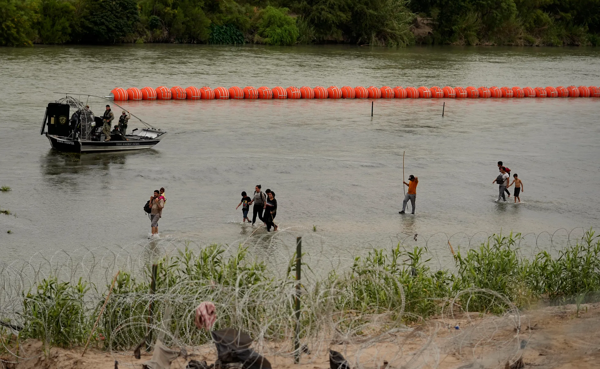 The buoy chain can be seen in the middle of the Rio Grande River while prospective immigrants wade in the river and a border patrol boat moves by