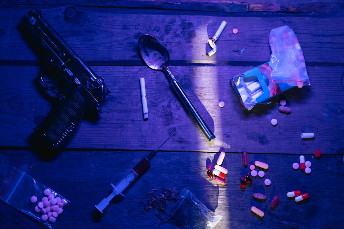 Items including pills, needles, spoons, rolled joints, and a pistol are strewn about a table