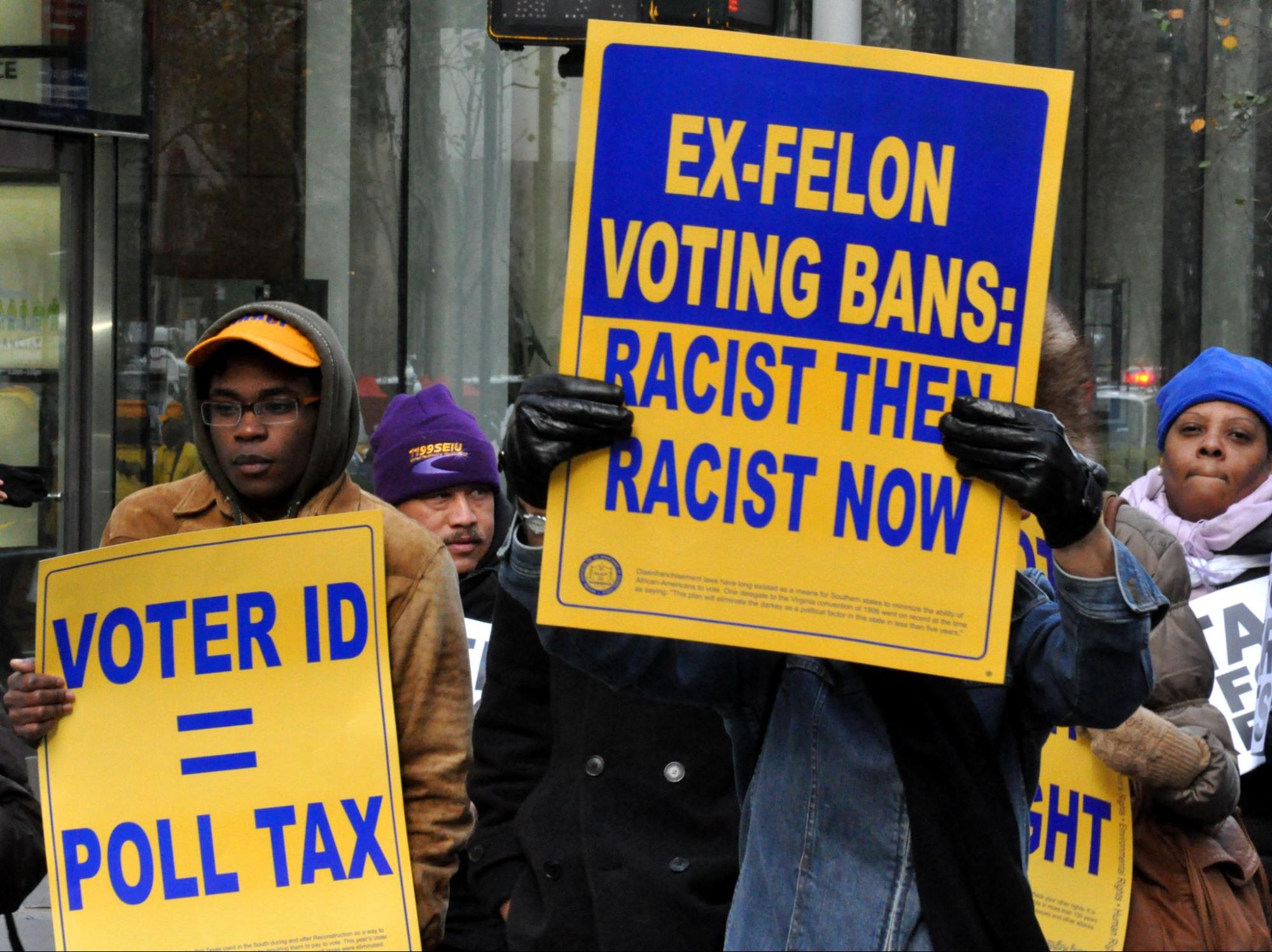 A group of individuals protesting in the street and holding signs that say, "ex-felon voting bans: racist then, racist now" and "voter ID = poll tax"