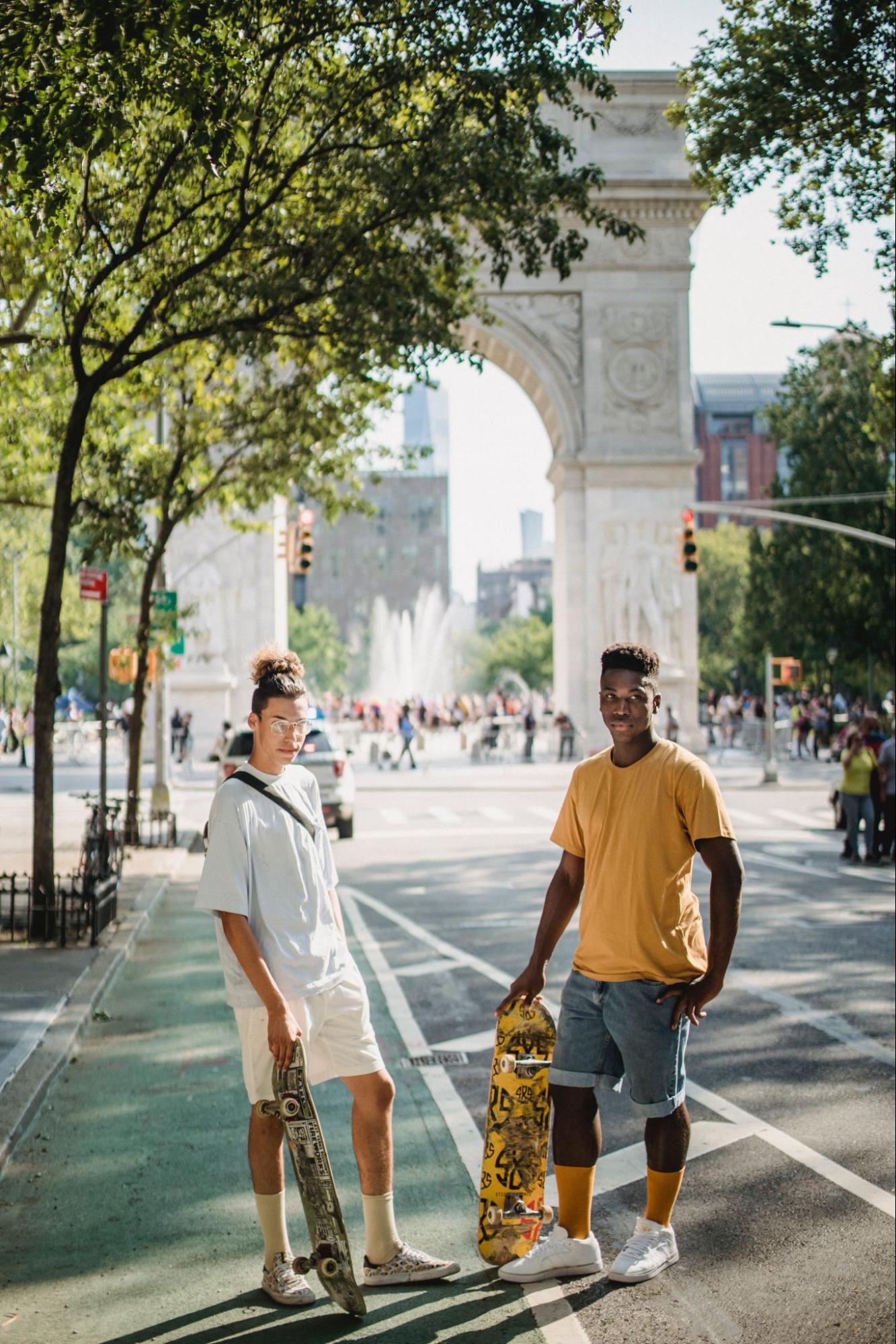 Two young men, one with light skin and one with dark, stand next to each other in a city holding skateboards