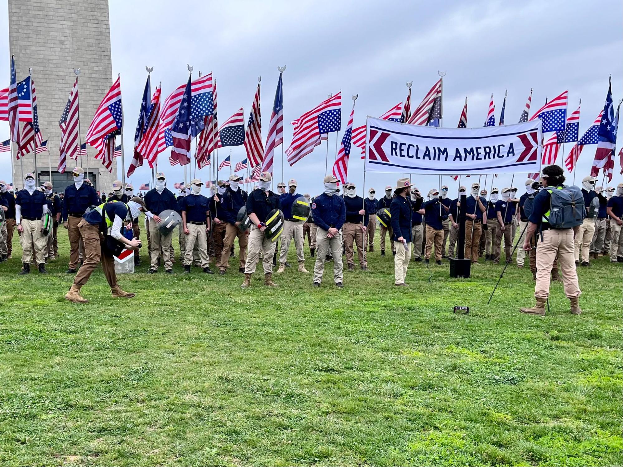 A group of people all wearing khaki pants, navy shirts, khaki ball caps, and face coverings. They are standing at attention and holding upside-down American flags while a sign that says "Reclaim America" is at the front of the group.