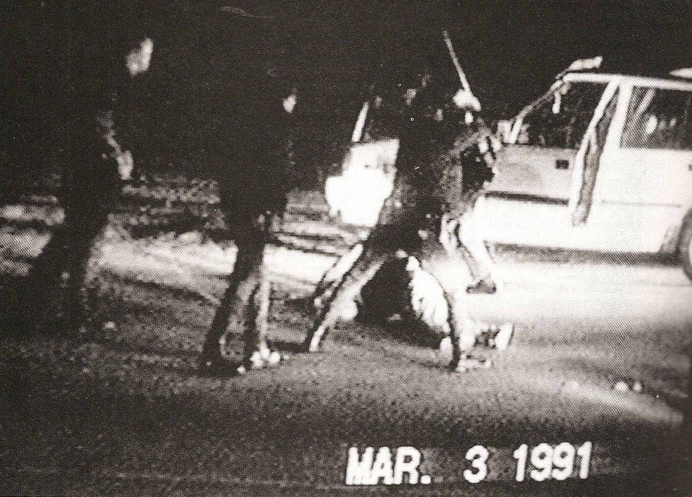 Black and white still shot from the famous video that captured police beating Rodney King. You can see three to four officers surrounding King who is on the ground, with a police vehicle in the background
