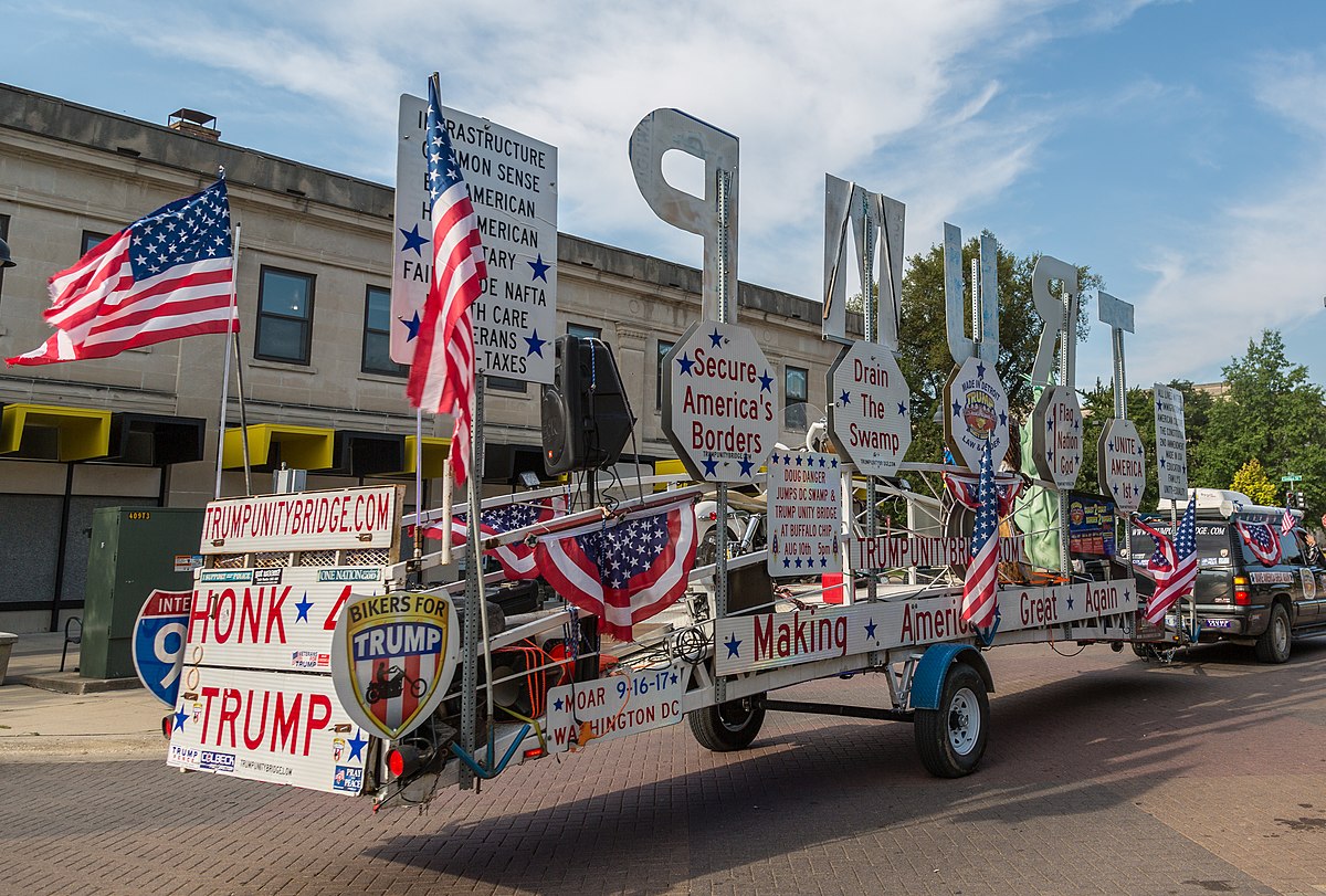 A trailer has been turned into a "float" for a "Trump parade." It has many American flags and signs with sayings such as "drain the swamp," "honk for Trump," and "Make America Great Again."