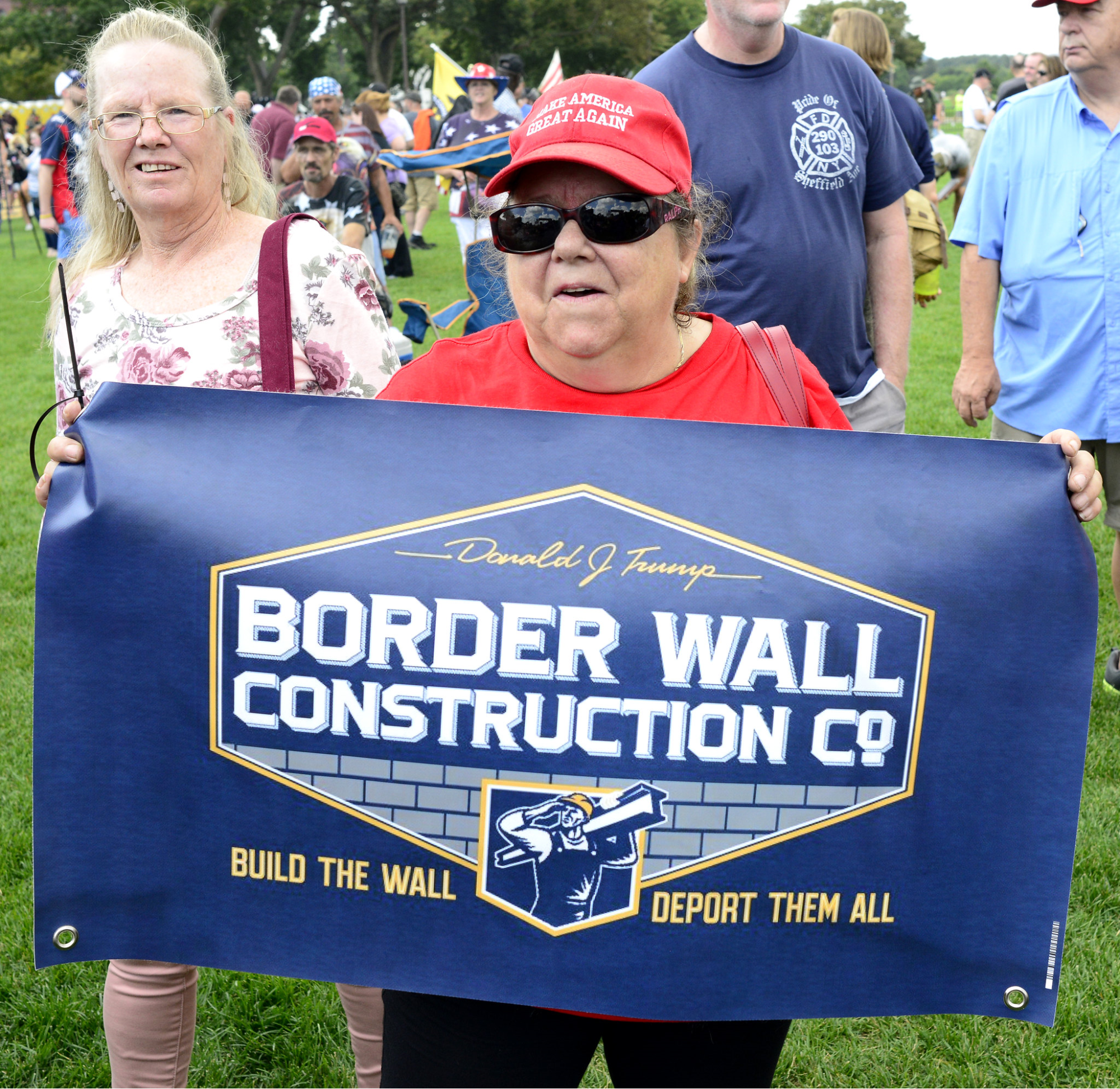At a Trump rally, a woman wearing a MAGA hat holds a sign that reads, "Donald J. Trump Border Wall Construction Co - Build the wall, Deport them all"
