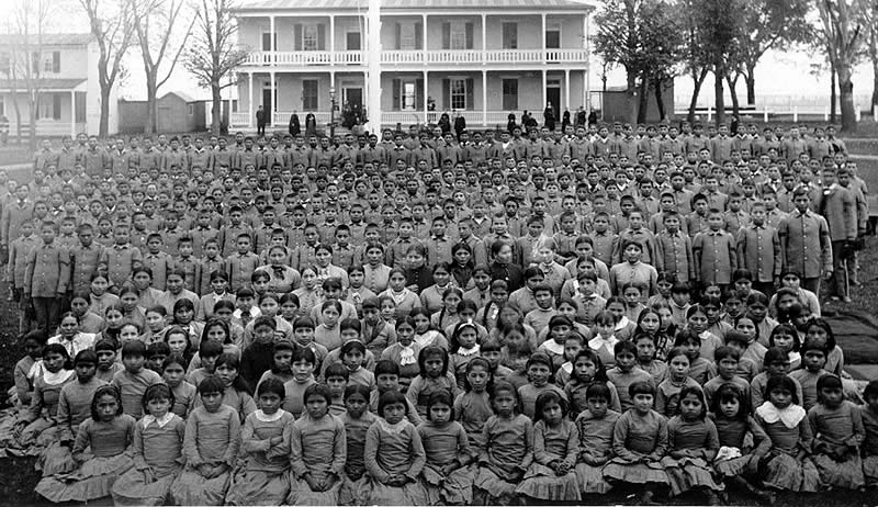 Black and white image of students at the Carlisle Native Industrial School in Pennsylvania (c. 1900) - there are so many children on the lawn in front of the "school" that you cannot even see the grass