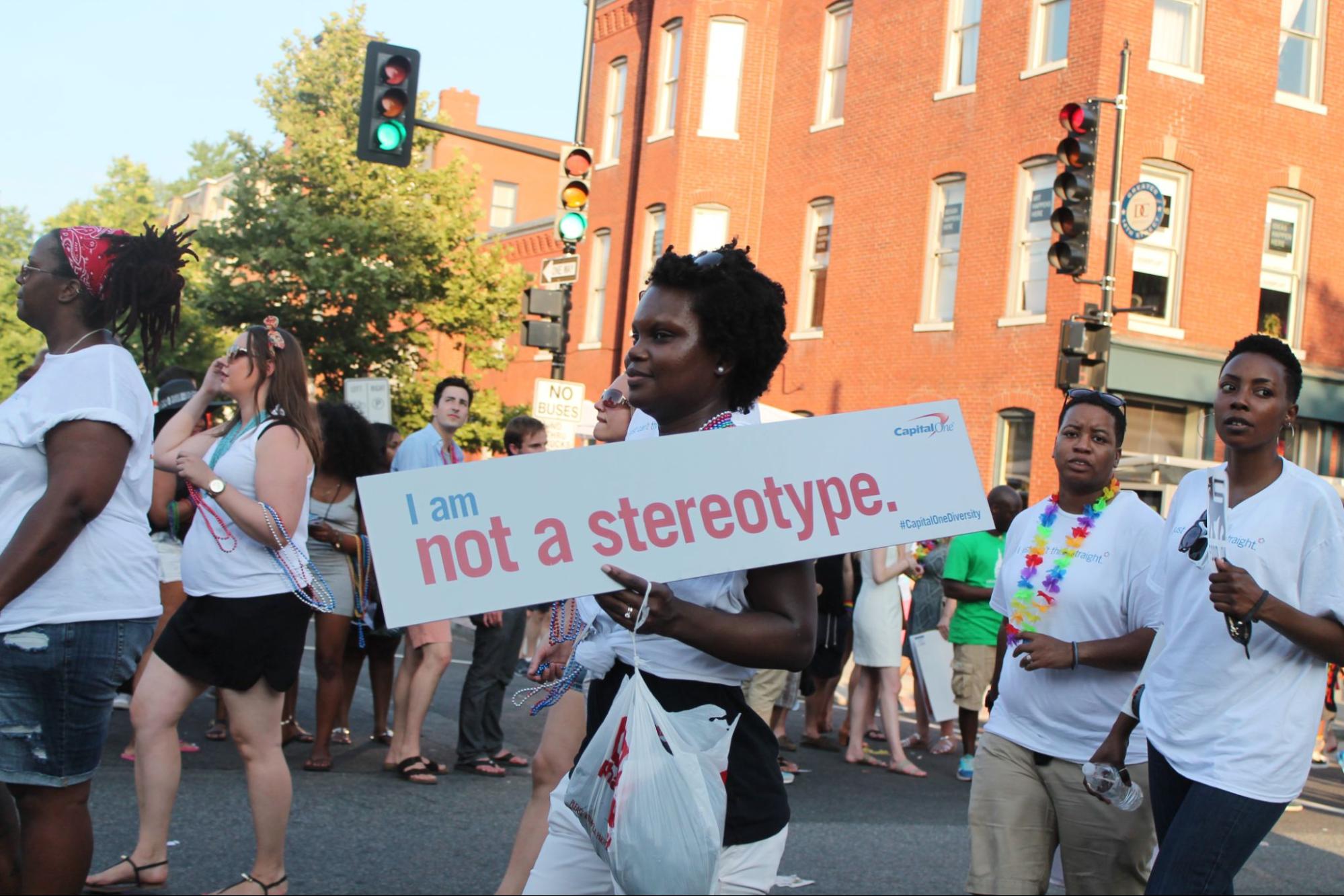 A Black woman walks down a street among other protestors with a sign that reads, "I am not a stereotype"