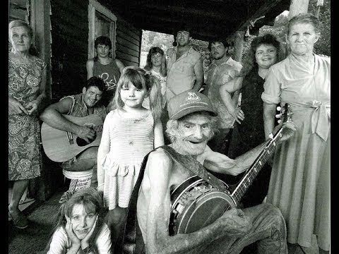 Black and white photo of a large white family gathered on the porch of their old home. Some are holding banjos or guitars.