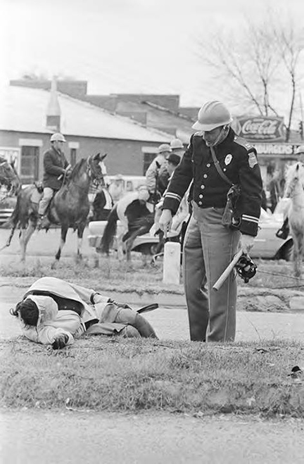 Black and white image of Amelia Boynton Robinson lying on the ground unconscious while a police officer stands over her and police on horseback can be seen in the background
