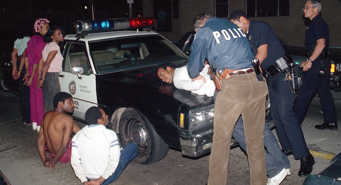 A police cruiser from the late 1980s is surrounded by Black individuals who have been placed in handcuffs. Two officers are actively arresting a Black man who they have bent over on the hood of the vehicle