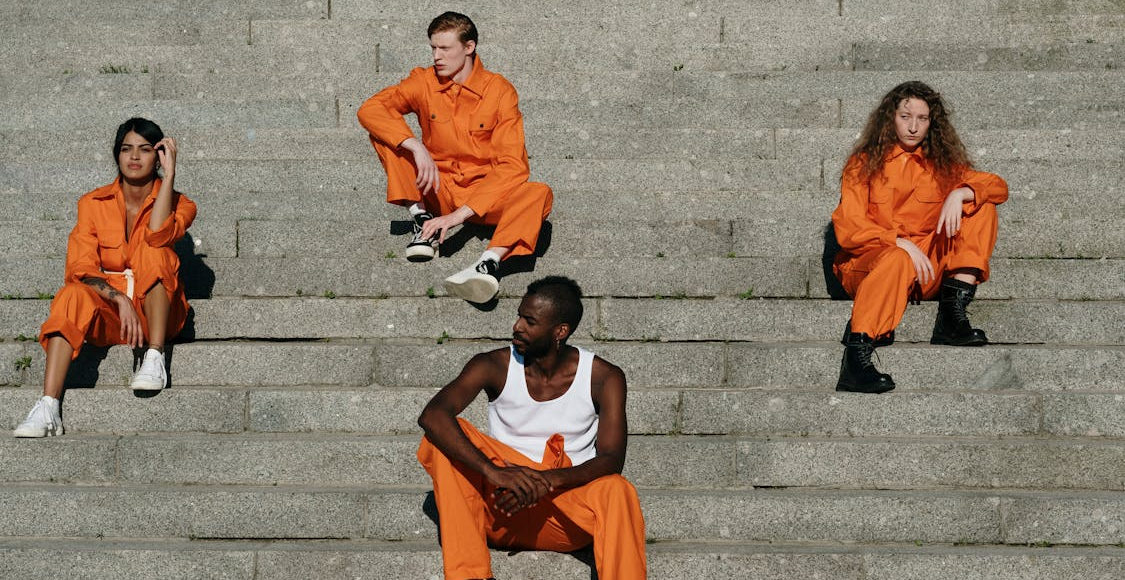 Four individuals of different genders and races sitting on steps and wearing orange prison jumpsuits