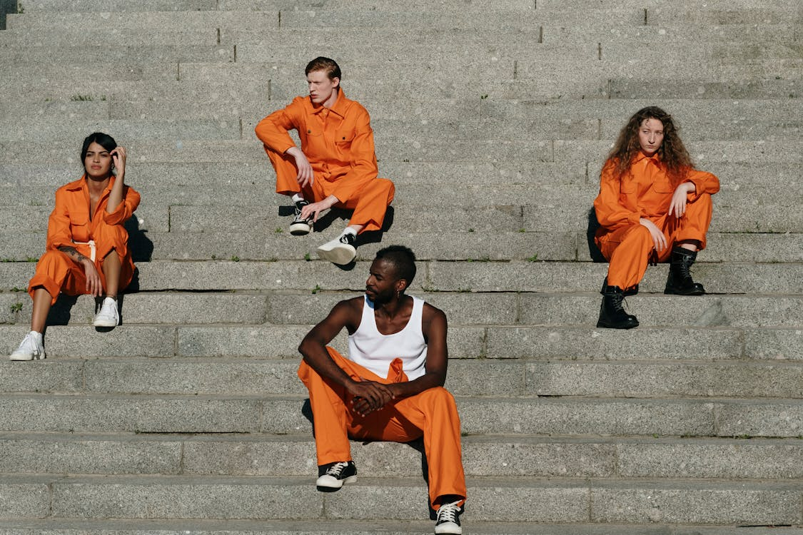 Four individuals of different genders and races sitting on steps and wearing orange prison jumpsuits