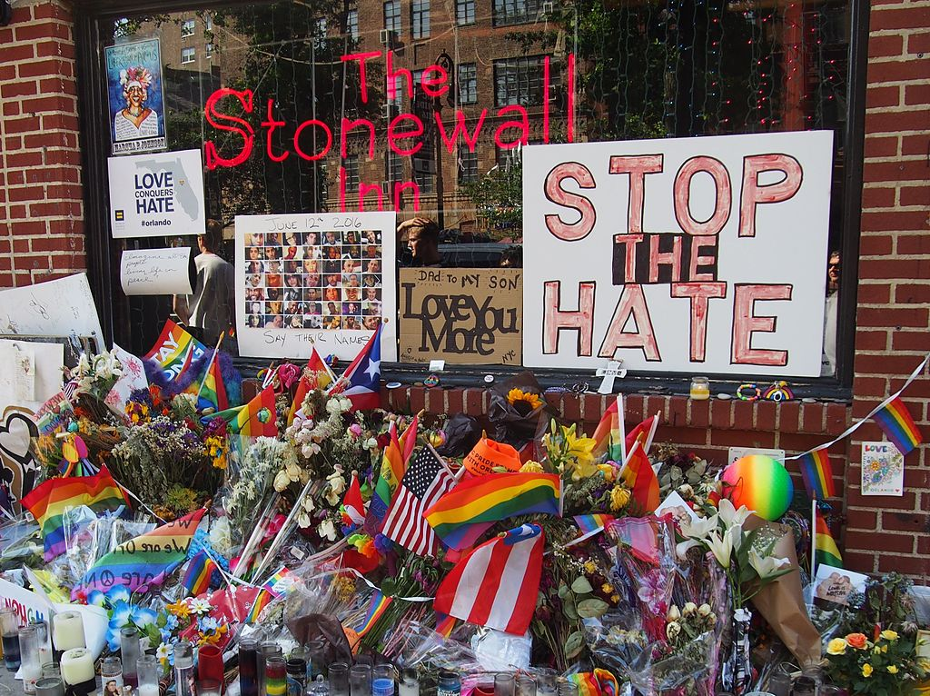 A memorial at the window of the old Stonewall Inn where individuals have placed signs such as "stop the hate" and various candles, Pride flags, and trinkets to honor the Stonewall Riot