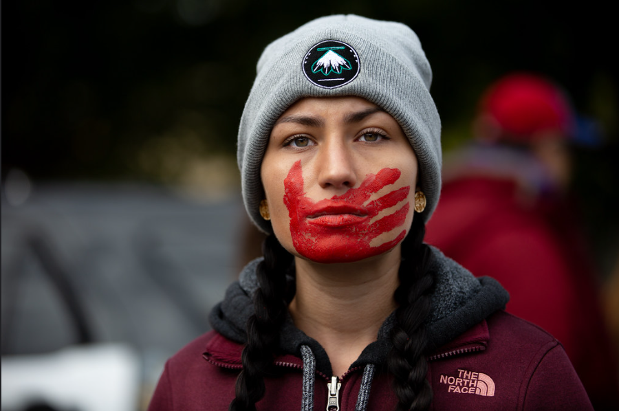 A person wearing a jacket and beanie with long dark braids looks at the camera with a red handprint painted over their mouth