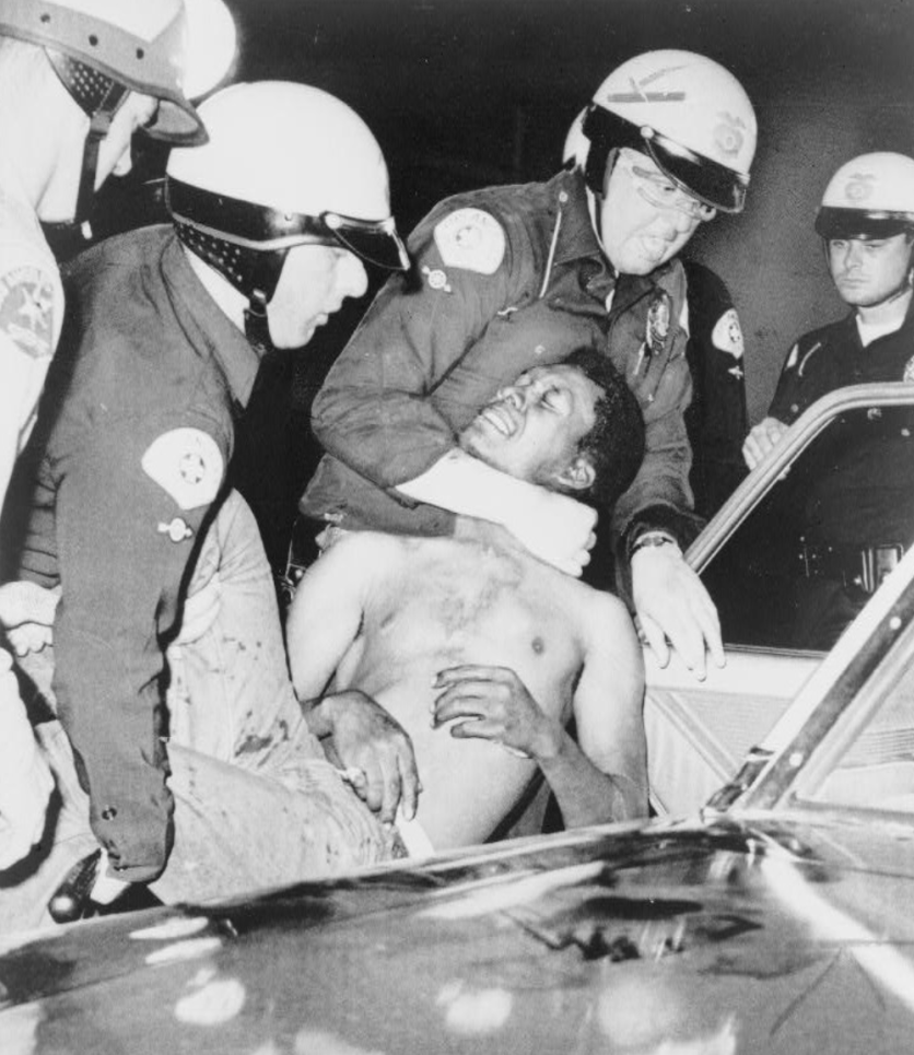Black and white image of four white police officers in helmets and goggles arresting and physically carrying - even by the neck - a young injured Black man.