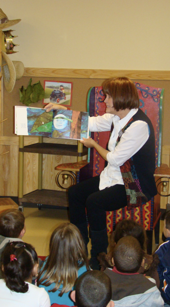 A woman sits in a chair holding and reading a book to children who are sitting on the ground in front of her.