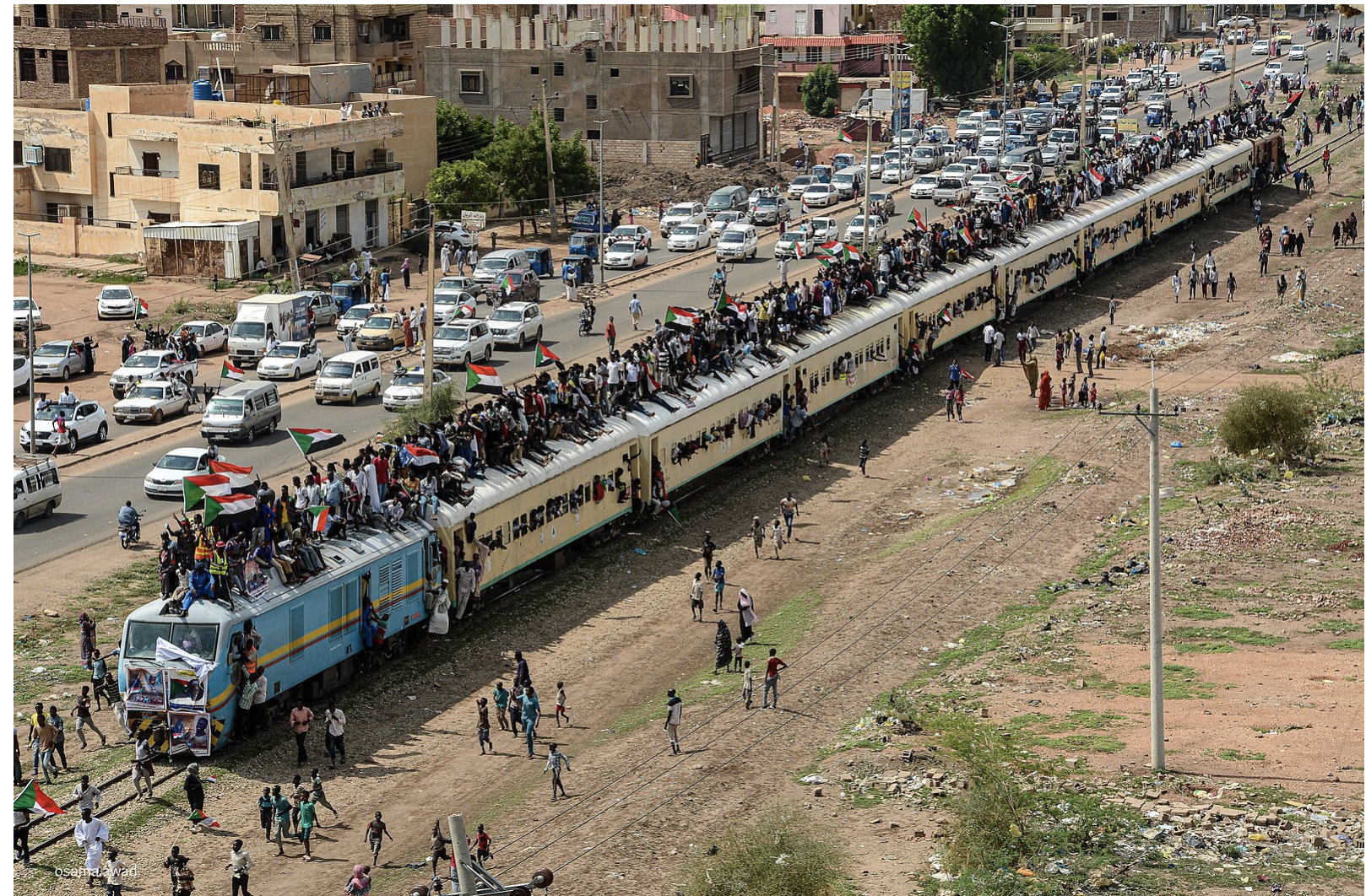 A train alongside a busy city road in Sudan is filled with people in and on top of the train in protest of genocide.