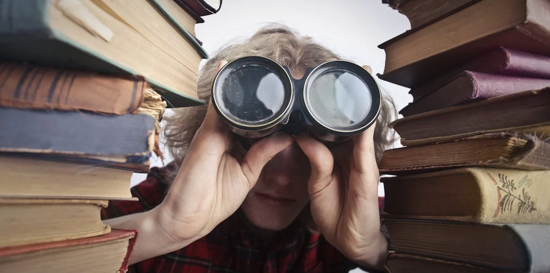 Between two tall stacks of old books, a person is leaning toward the camera peering into a large pair of binoculars