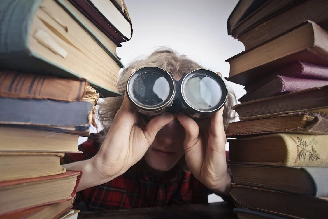 Between two tall stacks of old books, a person is leaning toward the camera peering into a large pair of binoculars