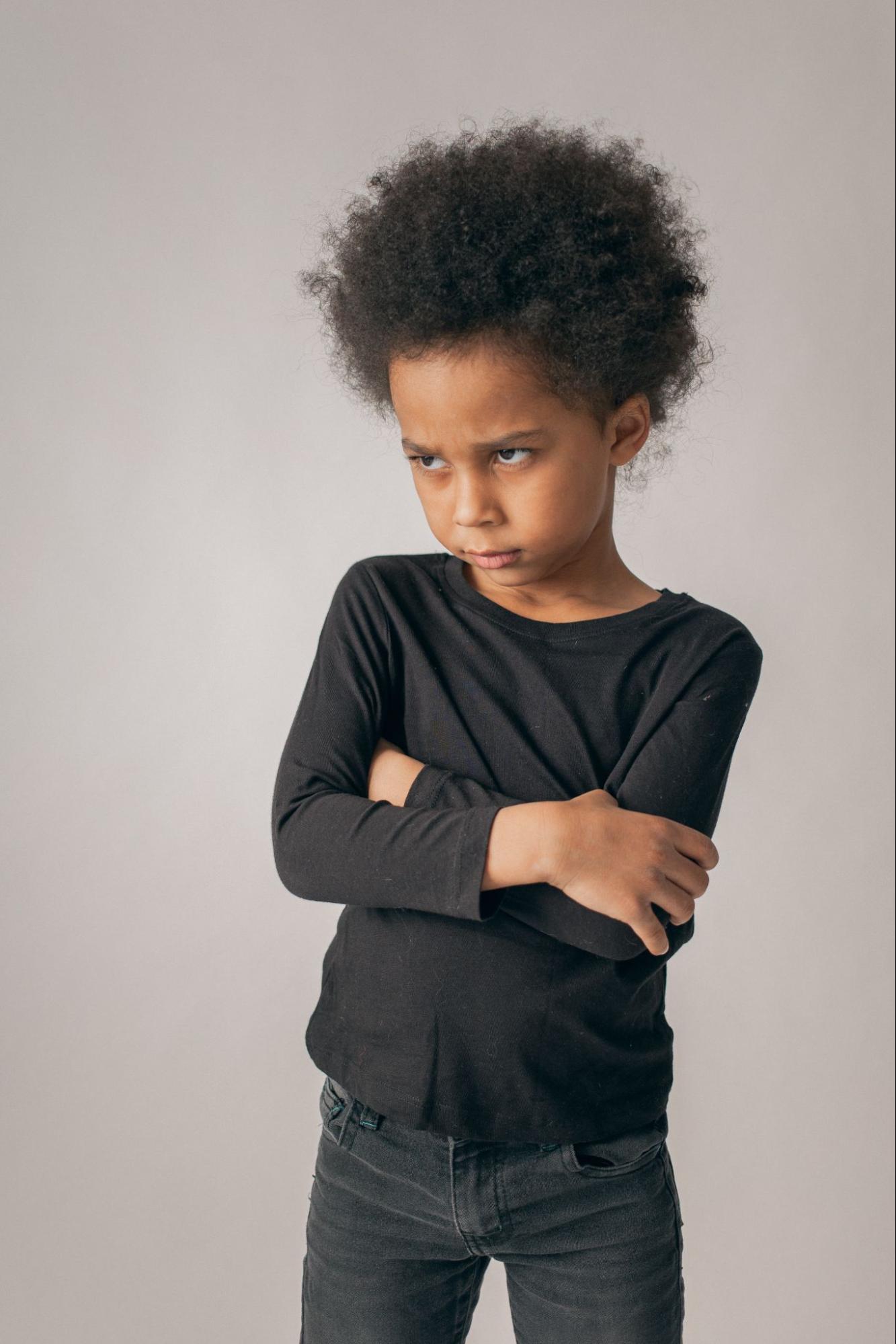 A Black child stands with their arms crossed and an upset look on their face