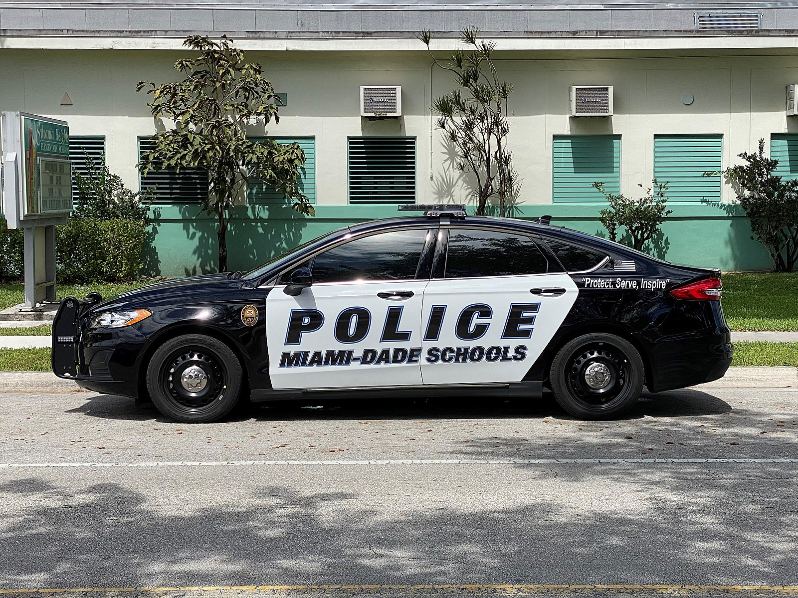 A police car in front of a school with the words, "Police Miami-Dade Schools" and "Protect, Serve, Inspire" on the side of the car